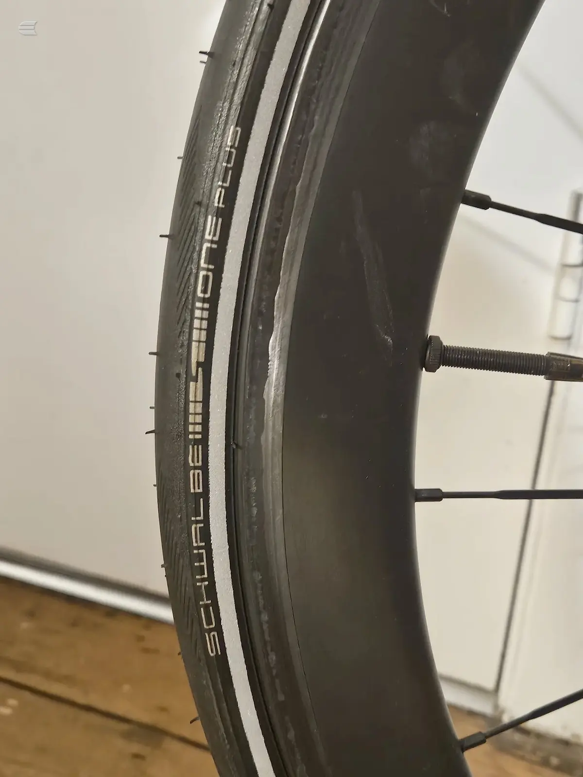 Close-up of a black Schwalbe bicycle tyre on a rim with white sidewall stripe, angled against a white wall and wooden floor, with brake cable visible.