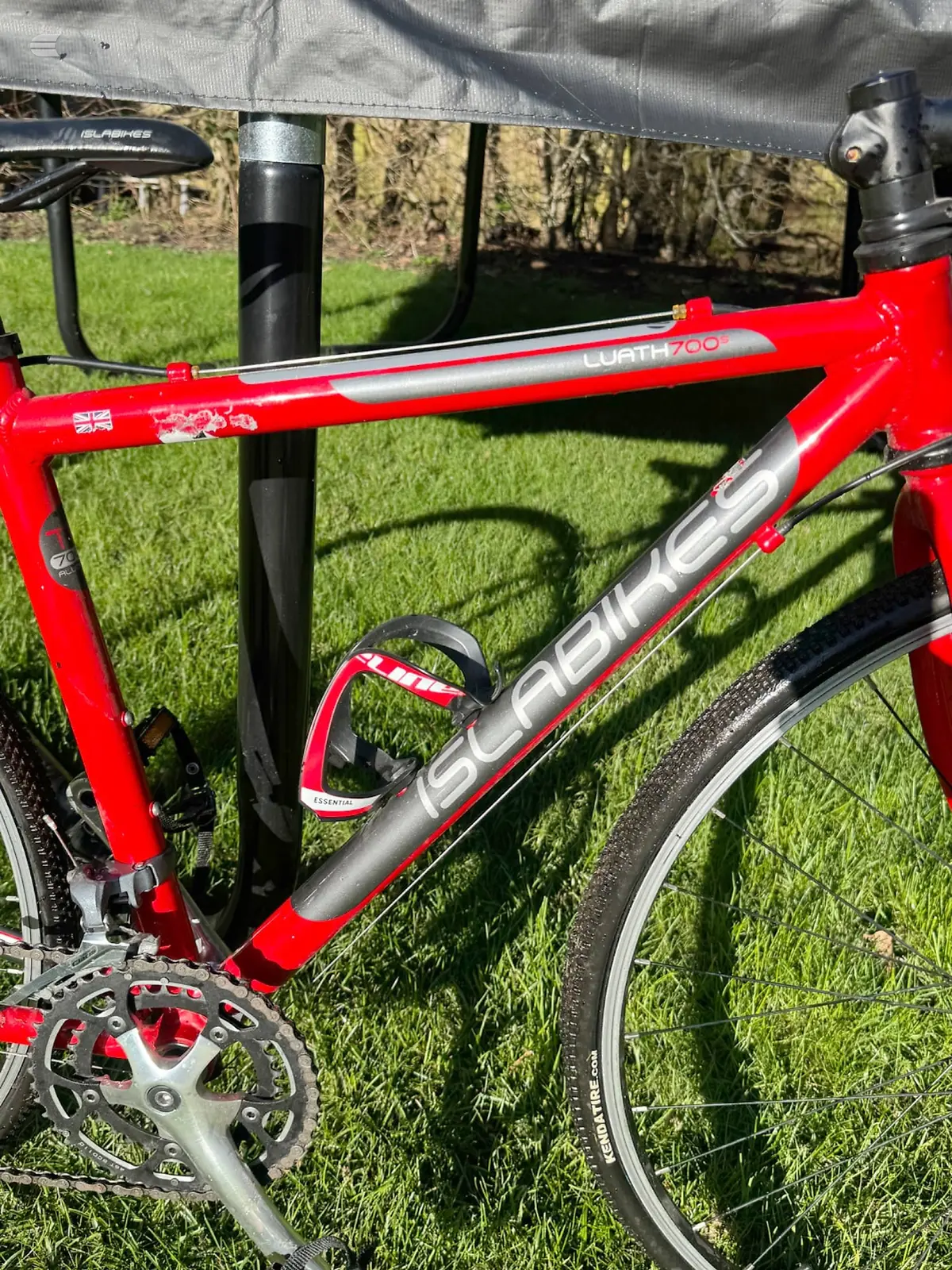 Red Scabreus road bike on grass, drive side, leaning against a black post with a bike rack visible in background.
