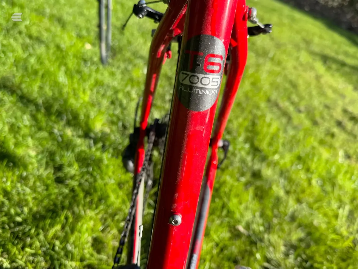 Red aluminium bicycle frame close-up showing head tube with '6' and '7005 ALUMINIUM' branding, on grass background, non-drive side angle.