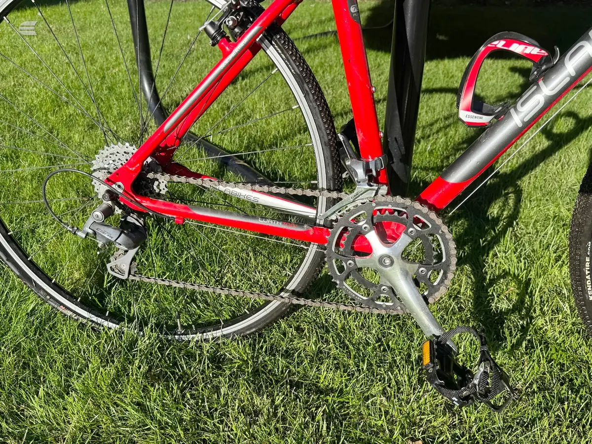 Red road bike drivetrain detail on grass, drive side, showing crankset, chain, and rear derailleur in sunlight.