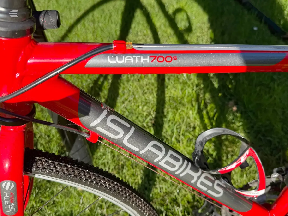 Close-up of red Wrath 700+ gravel bike frame and fork detail, drive side, on grass with shadow overlay.