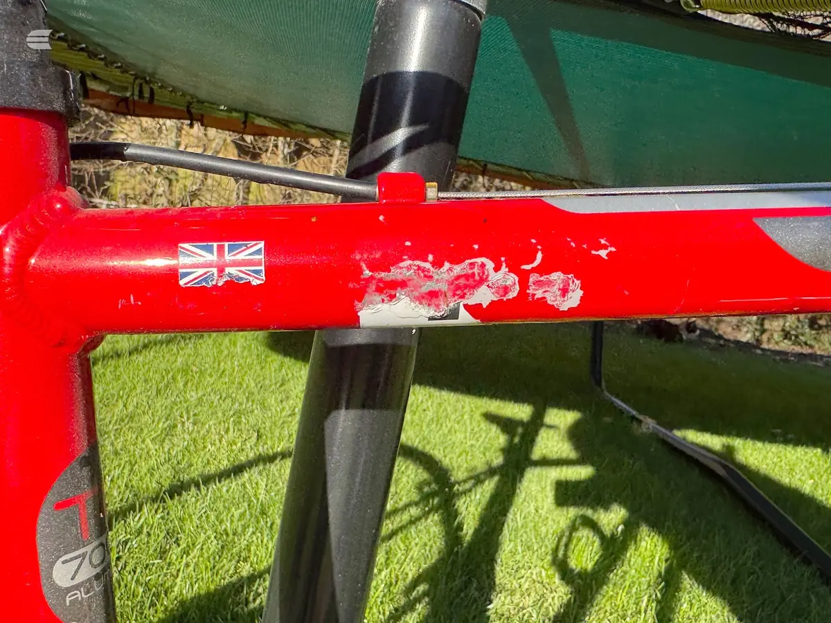 Red British-made bicycle frame with visible paint damage and wear on the top tube, black seat tube visible, green background.