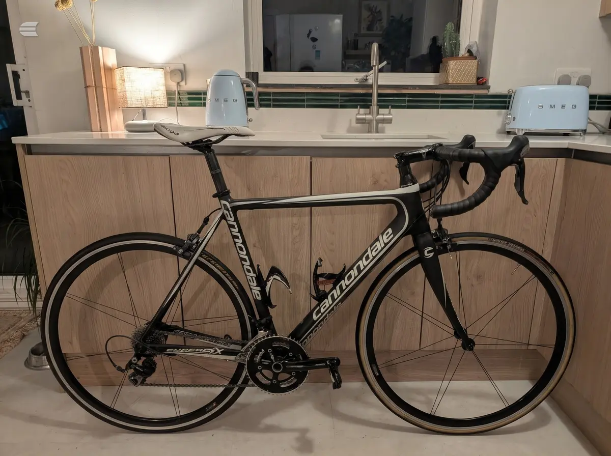 Black Cannondale road bike with drop bars, drive side, positioned in a modern kitchen against a wooden cabinet and white countertop.