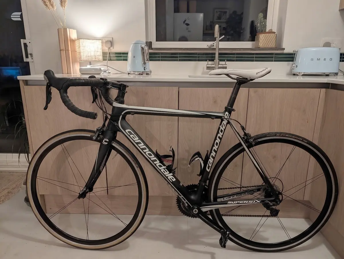 Black Cannondale road bike, drive side, indoors against a kitchen counter with appliances and window behind.