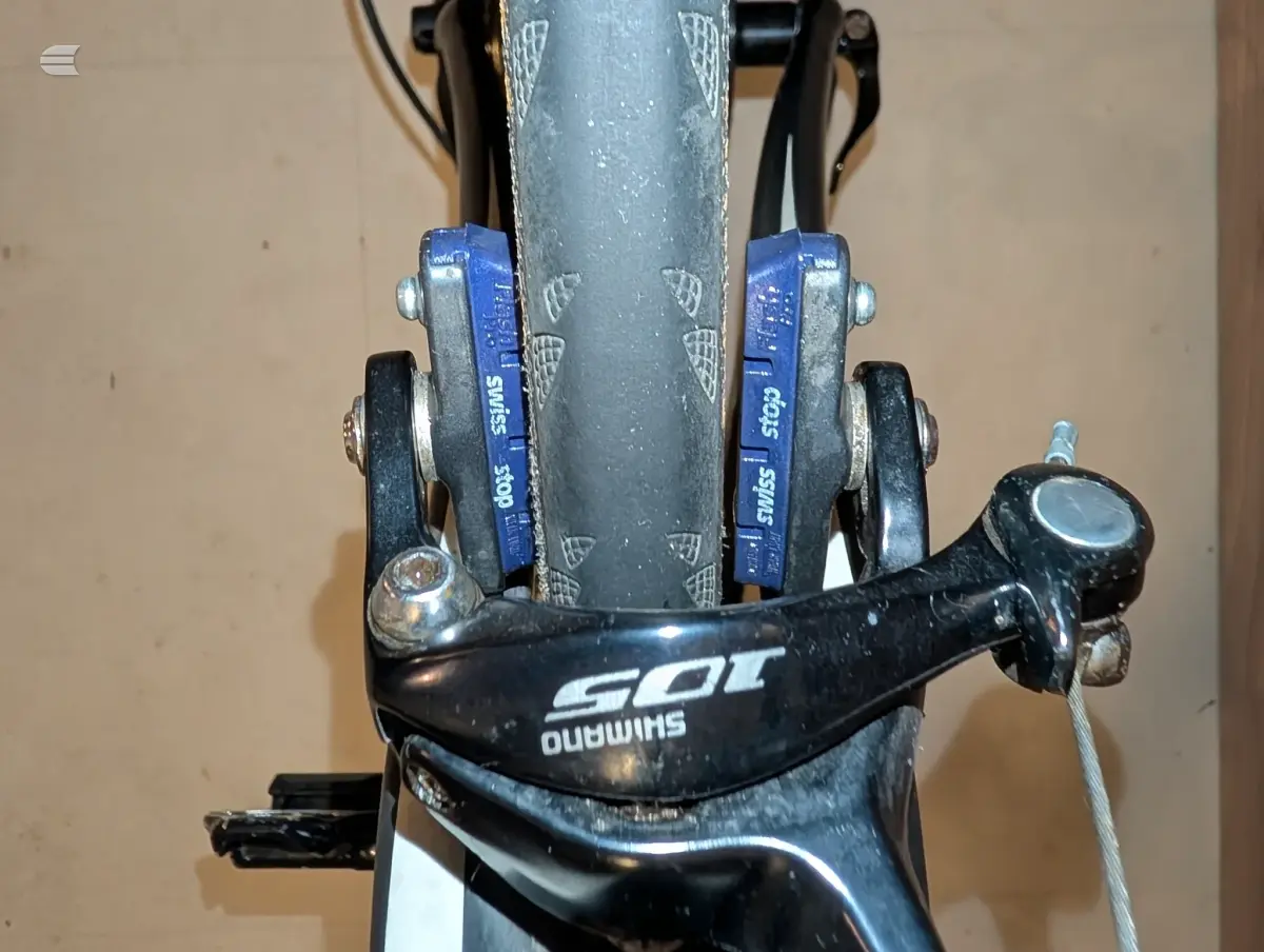 Close-up of Shimano 105 brake calipers on a bicycle with blue brake pads and grey rim, viewed head-on against a beige wall.