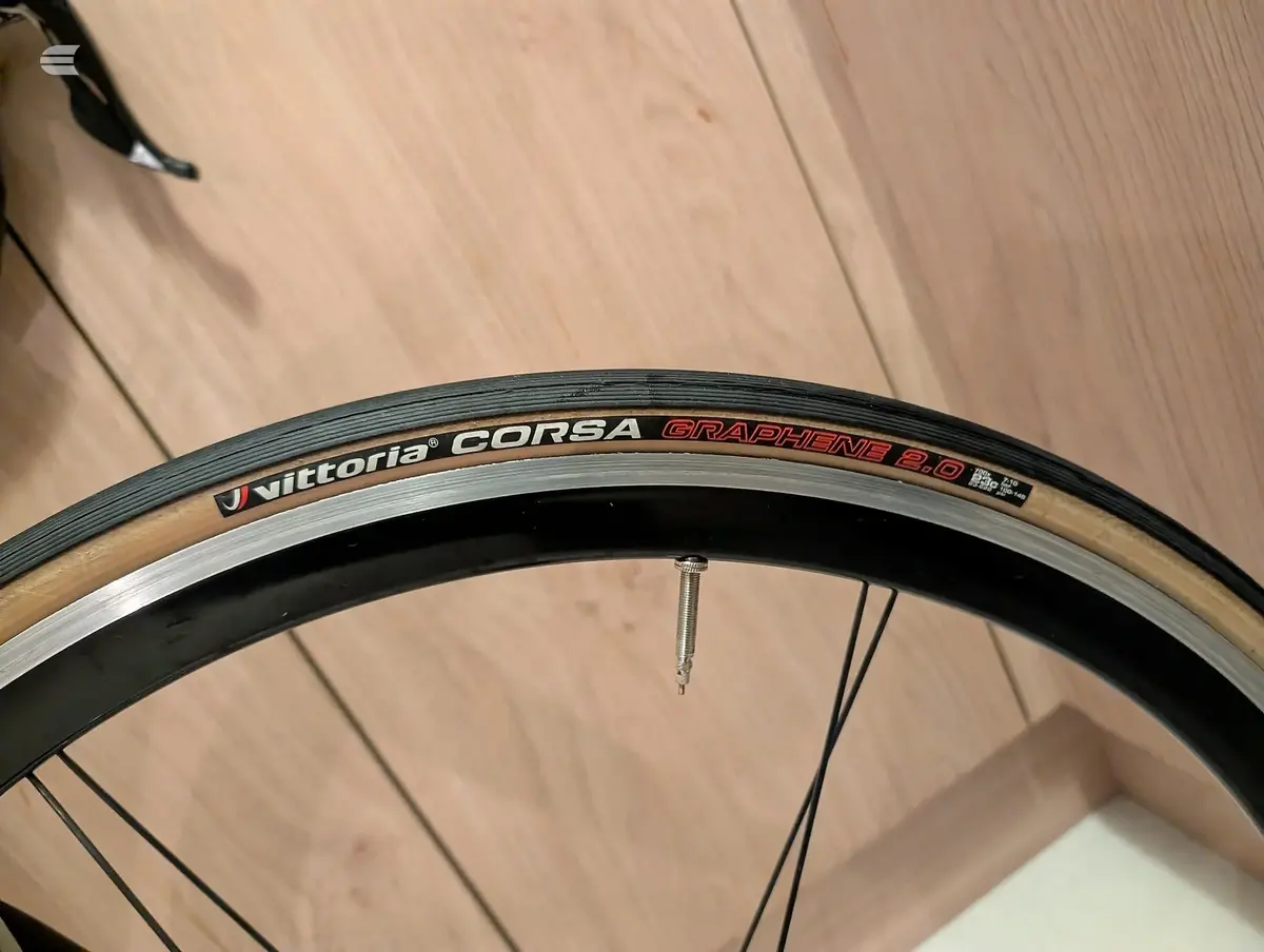 Close-up of a Vittoria Corsa Graphene 2.0 road bike tyre on a wooden floor, non-drive side, showing the tread and sidewall branding.