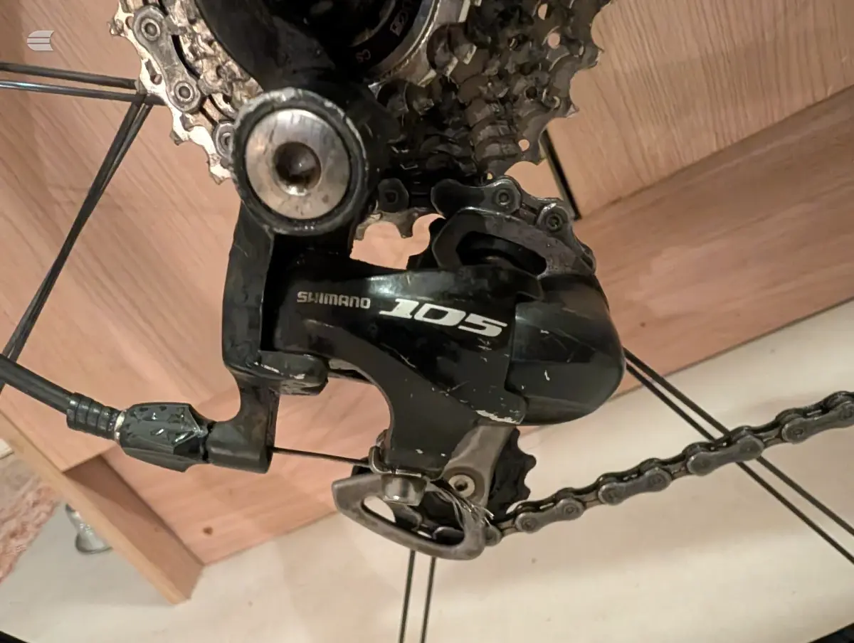 Shimano 105 rear derailleur on black bike, close-up from above showing pulley wheel and cage mechanism against wooden floor.