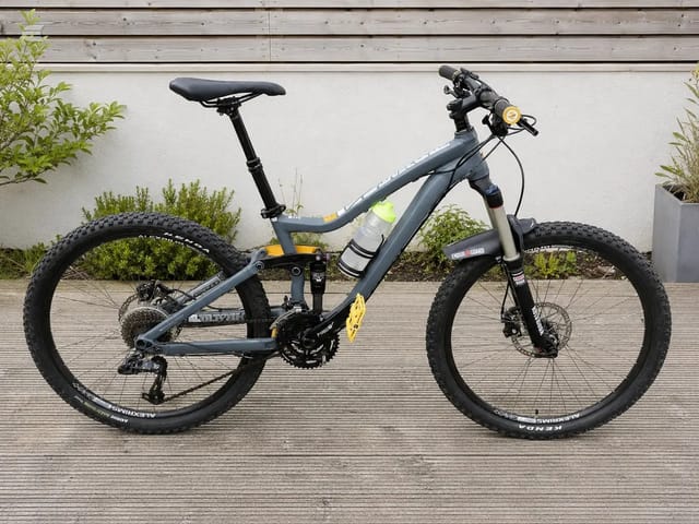 Grey and yellow full-suspension mountain bike on wooden decking, drive side, against white wall with potted plants.