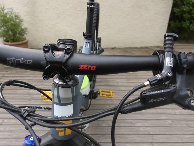 Close-up of mountain bike cockpit showing black Zeta handlebar with Strike stem, RockShox fork, and hydraulic disc brake lever, drive side, on wooden deck.
