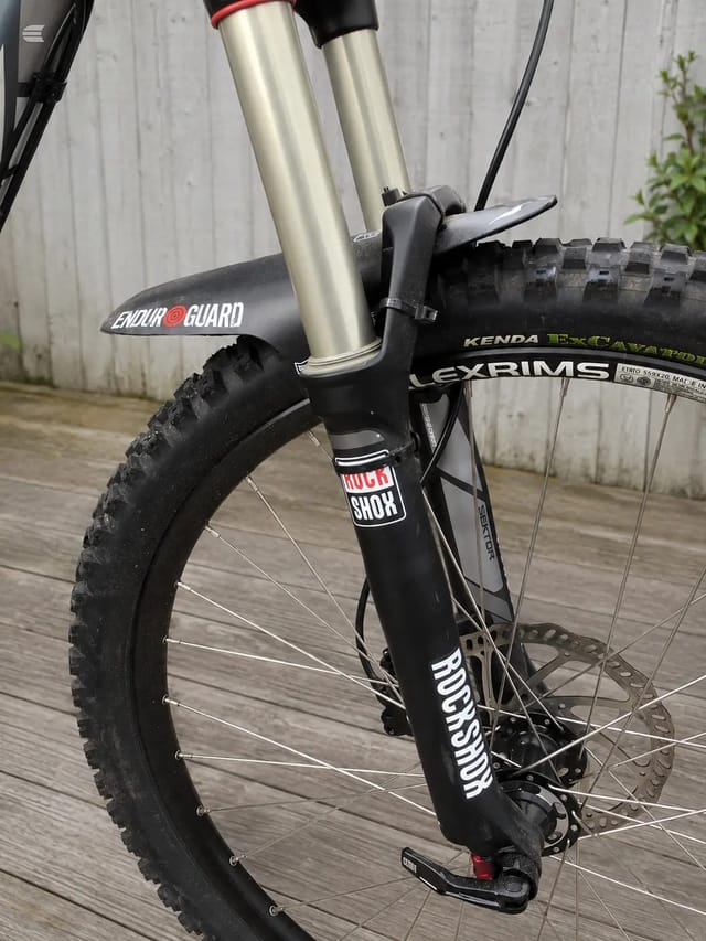 Black hardtail mountain bike with silver fork and red accents, front three-quarter view on wooden deck against concrete wall. Drive side visible.