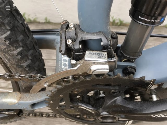 Close-up of mountain bike front derailleur and crankset, drive side, showing 9-speed SRAM component with chain and chainring detail against concrete background.