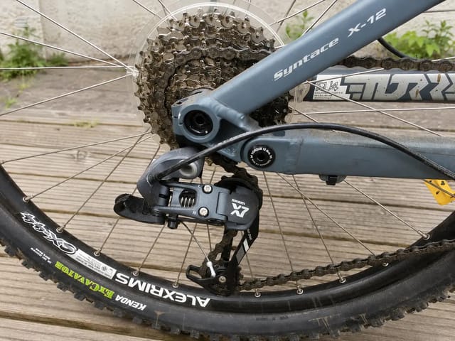 Close-up of SRAM X7 rear derailleur and Syntace X-12 seatstays on grey hardtail mountain bike, drive side, on wooden deck.