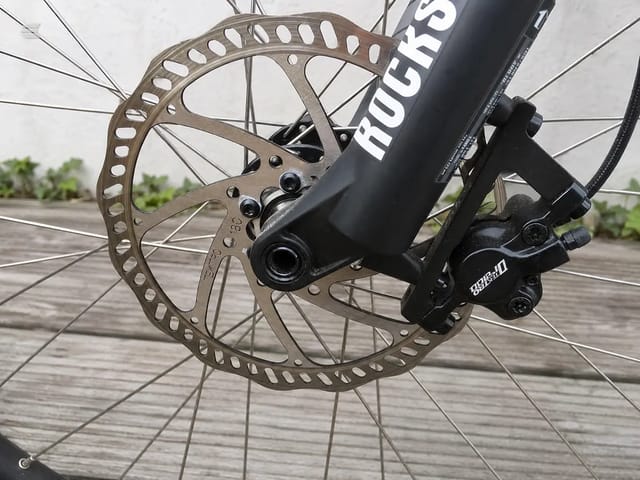 Close-up of hydraulic disc brake system on black mountain bike frame, front wheel with rotor visible, wooden deck setting, drive side angle.
