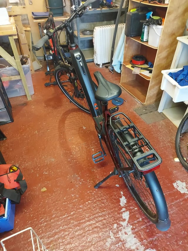 Black and red electric cargo bike on stand, drive side, in cluttered garage with wooden shelving and storage boxes.