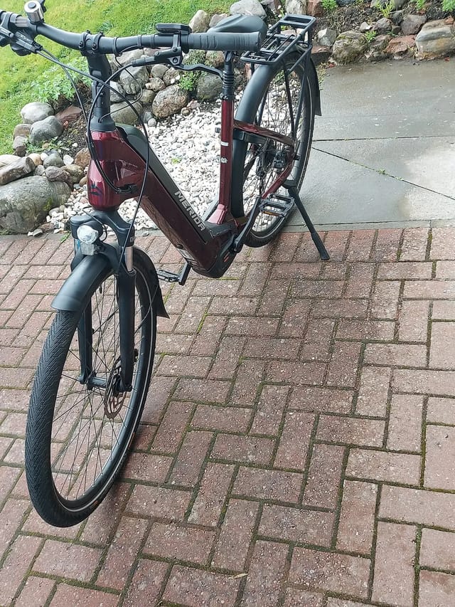 Burgundy and black step-through e-bike on a brick paved driveway, three-quarter view from the left side, with a residential garden and stone wall in the