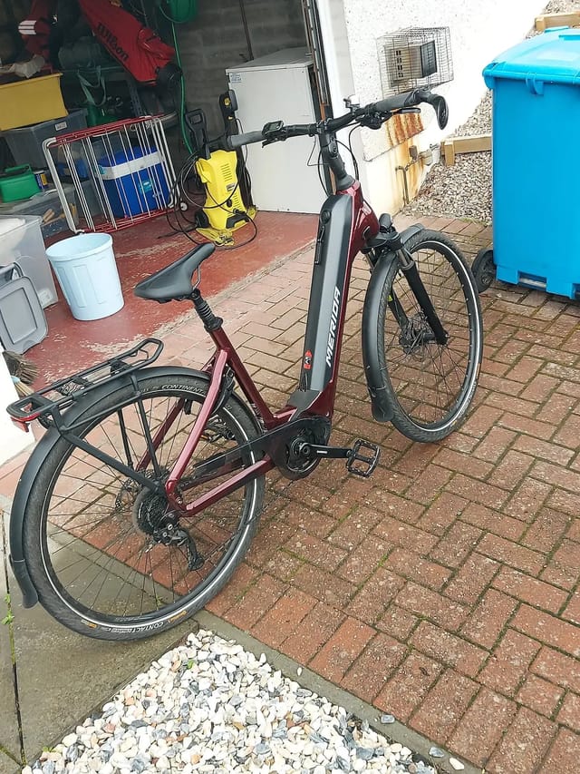 Maroon and black step-through e-bike, three-quarter view from the non-drive side, parked on a brick patio in a residential courtyard.