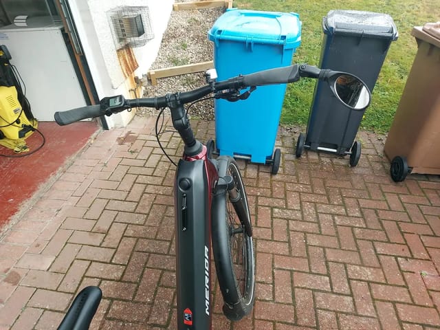 Black and maroon Merida gravel bike on brick paving, drive side, facing left, with blue and black wheelie bins and garden setting behind.