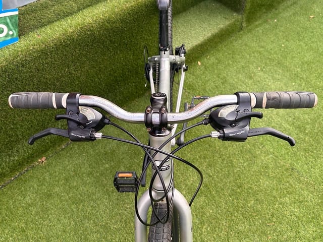 Silver hardtail mountain bike handlebars and stem, front-on view on artificial grass. Tektro brake levers and grips visible.