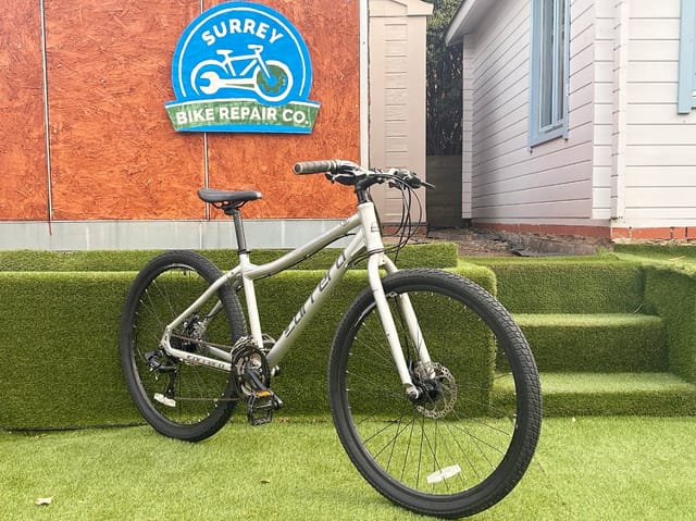 Silver hardtail mountain bike on grass, drive side, in front of Surrey Bike Repair Co storefront with orange plywood wall.