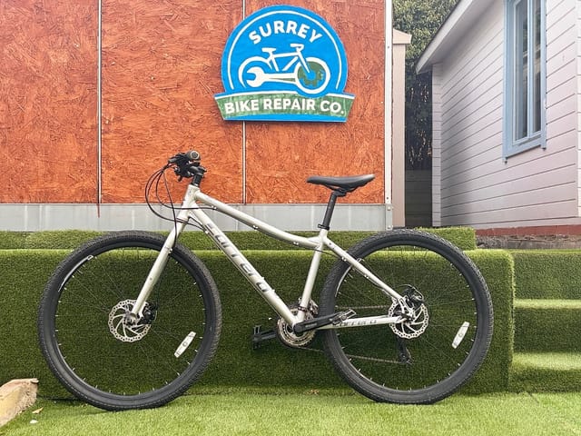 Cream hardtail mountain bike with disc brakes, drive side, on artificial grass in front of Surrey Bike Repair Co. storefront.