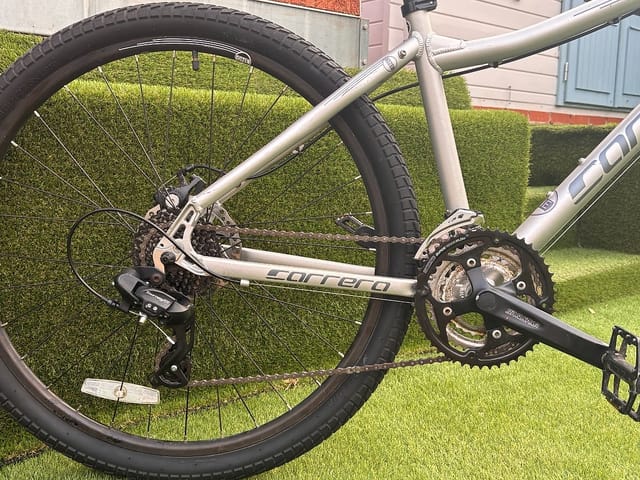 Silver Corratec hybrid bike on grass, drive side, with black tyres and dark drivetrain components visible.