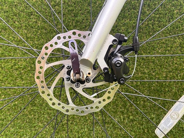 Close-up of disc brake system on white hardtail mountain bike, front wheel and brake caliper visible against green grass background.