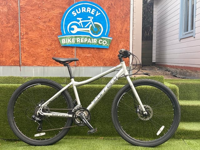Cream Cannondale hybrid bike on artificial grass, drive side, in front of Surrey Bike Repair Co. signage on orange plywood wall.
