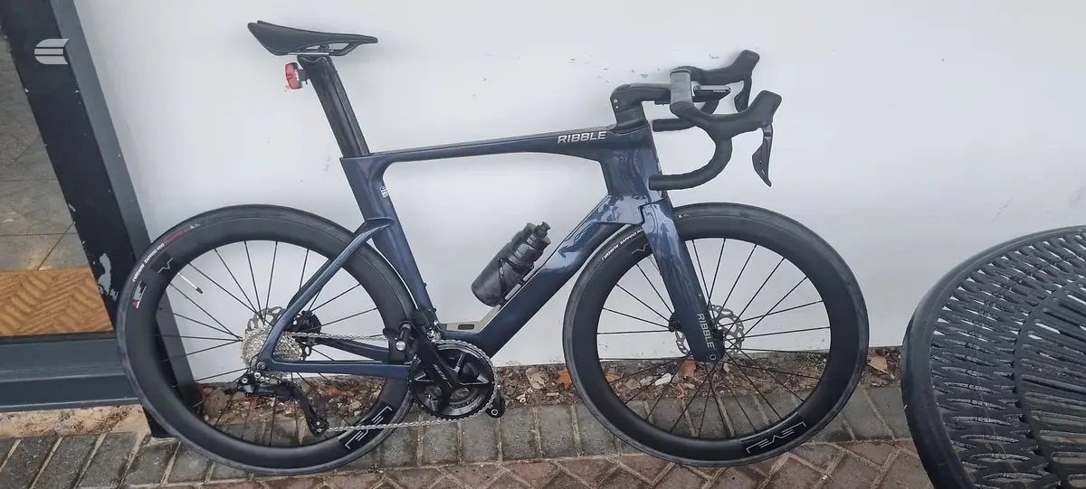 Dark blue Ribble aero road bike with disc brakes, drive side, parked against a white wall and brick base on a residential street.