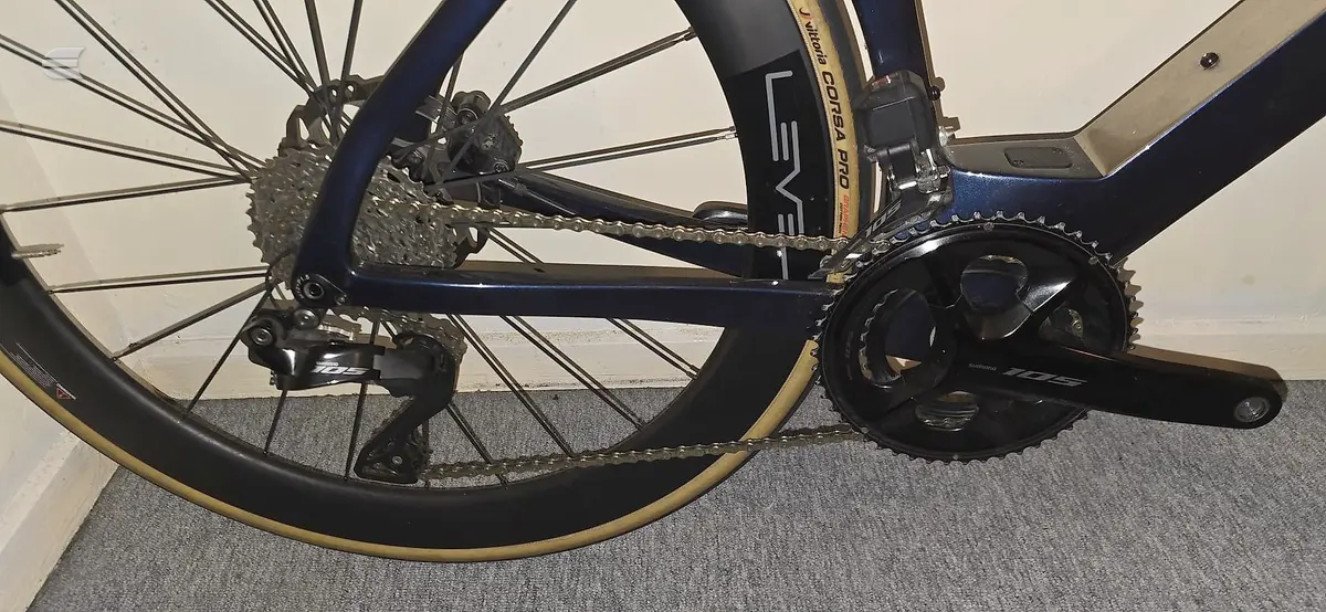 Dark blue road bike drivetrain detail, drive side, on grey carpet indoors. Close-up of crankset, chain, and rear cassette.