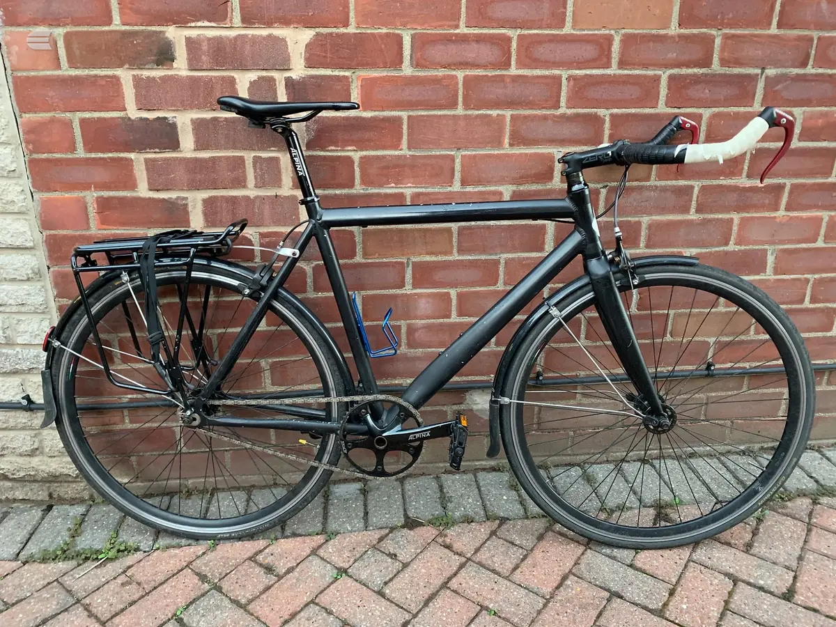 Black commuter bike with drop bars, mudguards, and rear rack, parked against a red brick wall, drive side view.