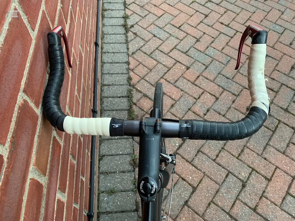 Black drop-bar road bike viewed from above, non-drive side, on herringbone brick paving next to a red brick wall.