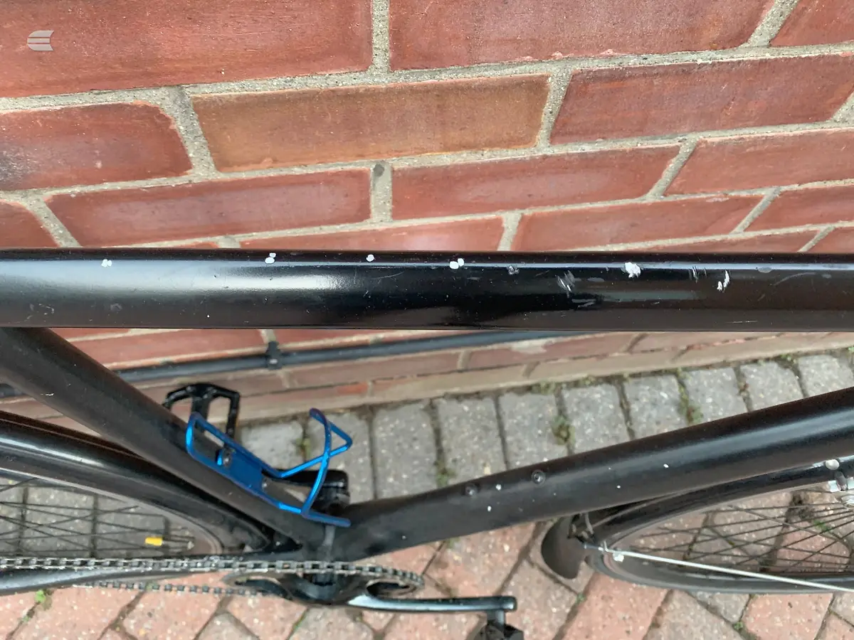 Black hardtail mountain bike with blue bottle cage, leaning against red brick wall, drive side view, partial frame visible with front wheel and handlebars in