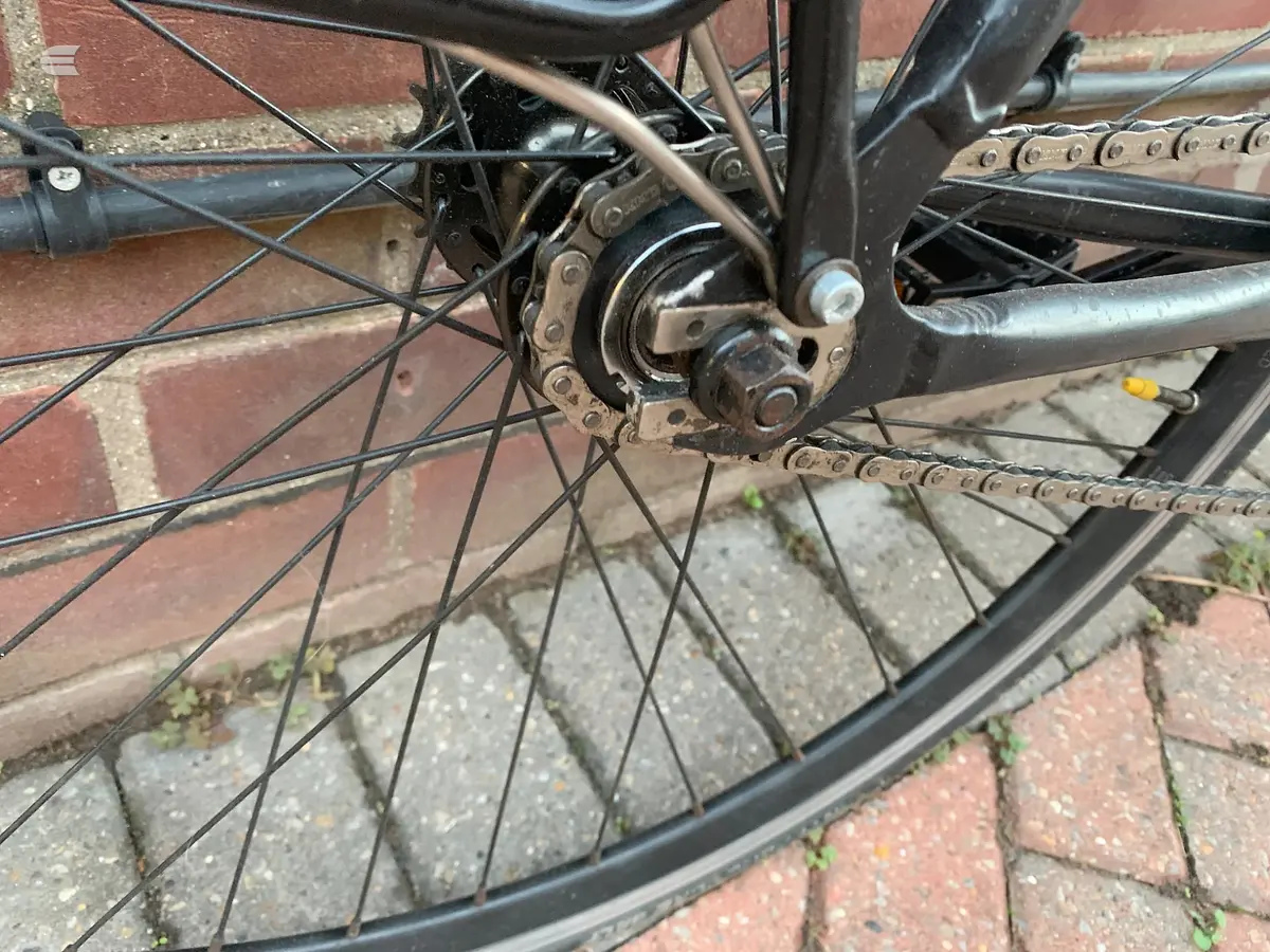 Close-up of bicycle rear hub, chain, and derailleur on dark frame, drive side, against brick pavement.
