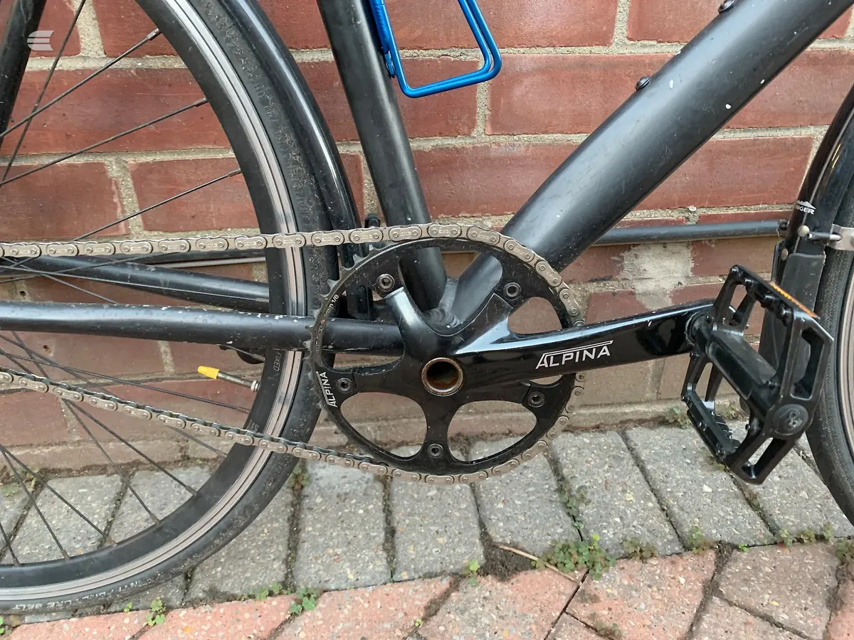 Alpina road bike crankset and chain detail, drive side, against brick wall and paving. Close-up of black crank arm with chainring.