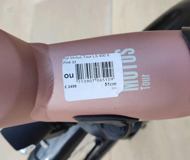Close-up of a price label on pink fabric, showing barcode, product code, price £24.99, size 51cm, and partial text 'Motus Tour LS 400' and 'Pink 22'.