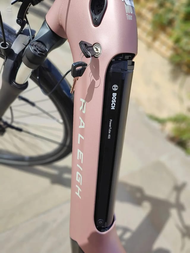 Pink Raleigh gravel bike, close-up of fork and seatpost with Bosch eBike motor and battery, non-drive side, on pavement.
