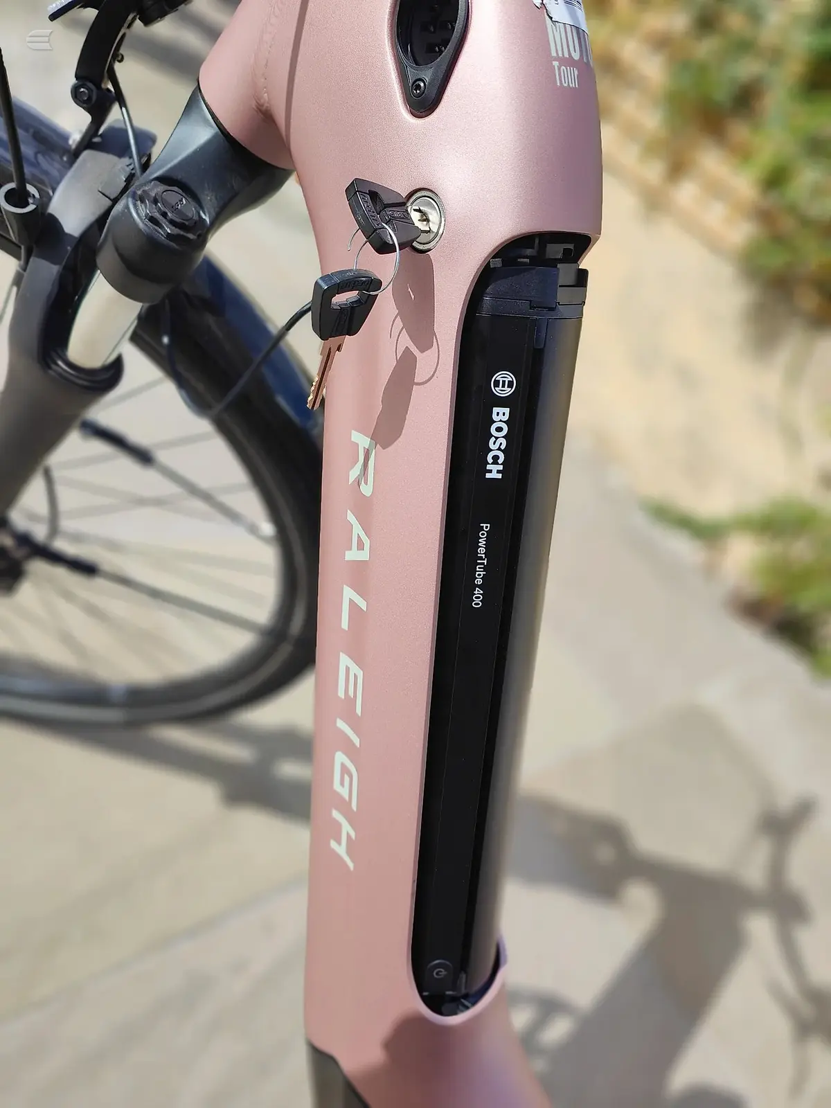 Pink Raleigh gravel bike, close-up of fork and seatpost with Bosch eBike motor and battery, non-drive side, on pavement.