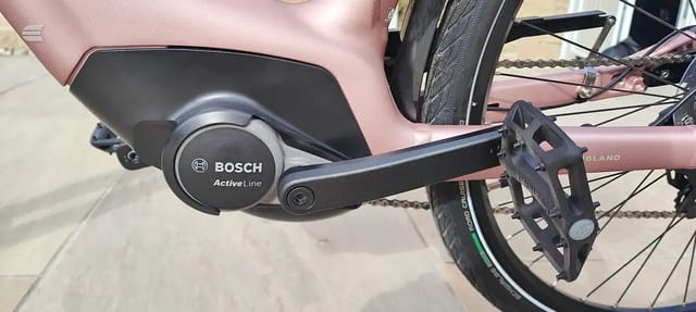 Bosch ActiveLine mid-drive motor and pedals on a pink e-bike, drive side close-up, outdoors on pavement.