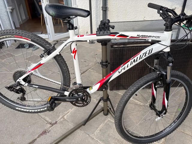 White and red Specialized Hardrock mountain bike on a stand, drive side, outdoors on pavement with a building and shutter behind.