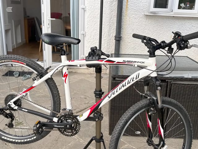 White and red Specialized Hardrock mountain bike on a repair stand, drive side, in a driveway with a house and grey render wall behind.