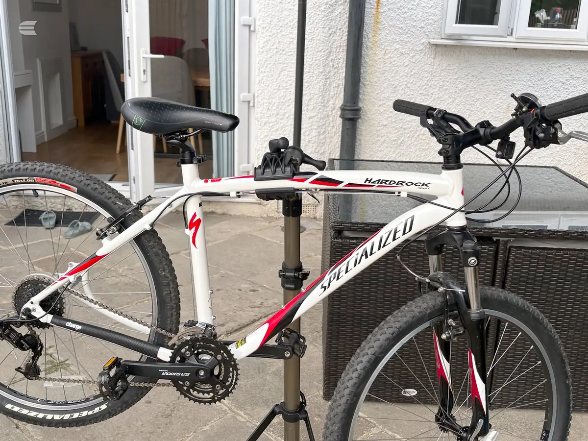 White and red Specialized Hardrock mountain bike on a repair stand, drive side, in a driveway with a house and grey render wall behind.