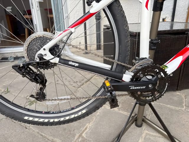 Red and white hybrid bike on a stand, drive side, showing chain, cassette, derailleur and crankset detail. Outdoor setting against a building.