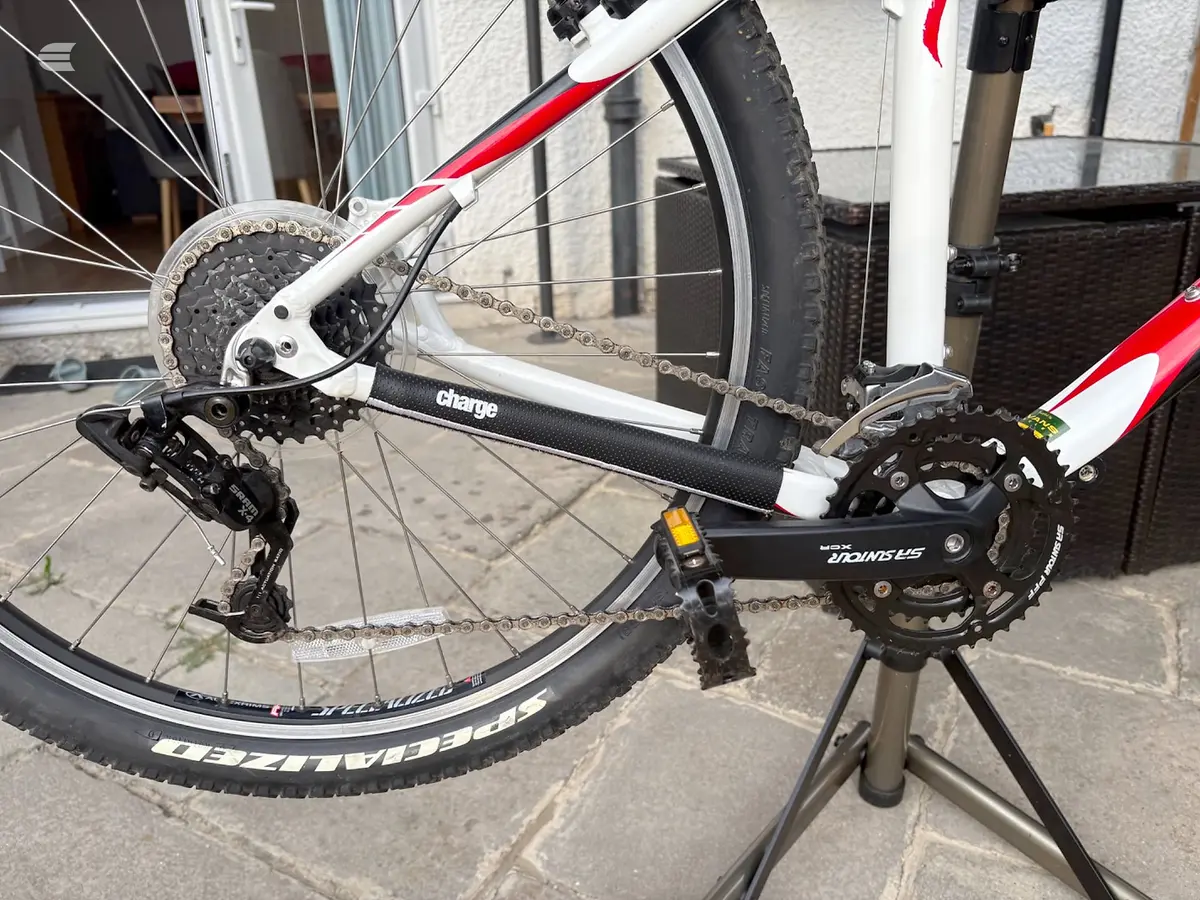 Red and white hybrid bike on a stand, drive side, showing chain, cassette, derailleur and crankset detail. Outdoor setting against a building.