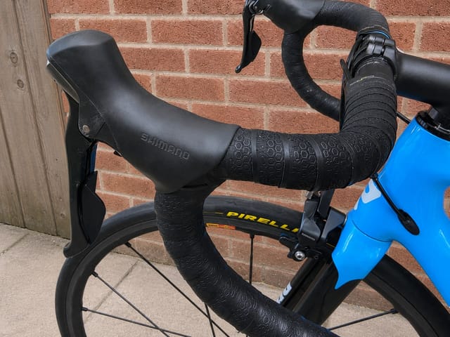 Shimano drop bar shifters and brake levers on black bar tape, blue road bike frame visible, against brick wall.