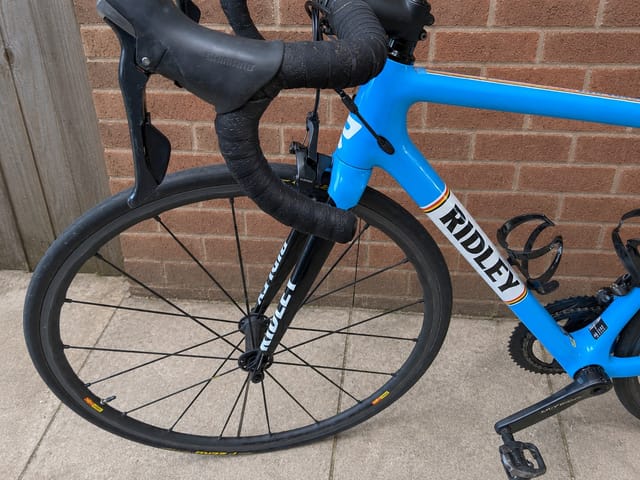 Blue Ridley road bike with drop bars, drive side, leaning against a brick wall on pavement.