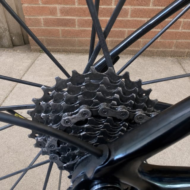 Close-up of a bicycle cassette and chain on the rear wheel, drive side, against a brick wall and pavement background.