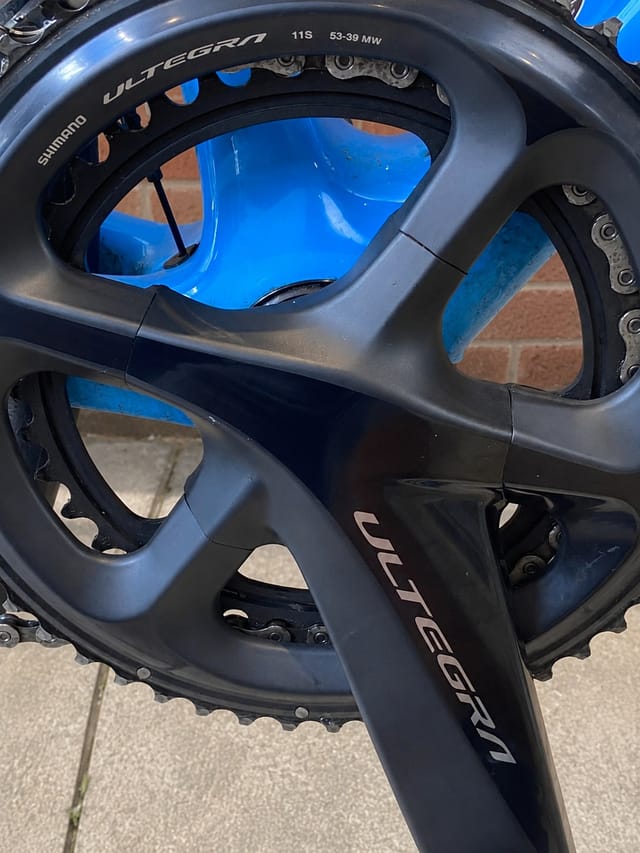 Close-up of Shimano Ultegra crankset and chainrings, drive side, against blue background and brick wall.