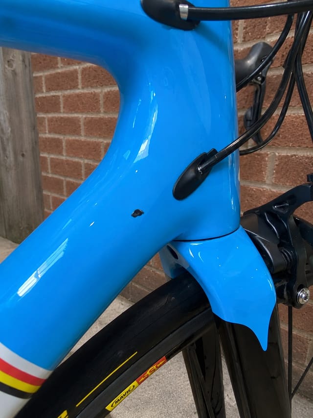 Blue road bike frame with visible chip or crack on seat tube, non-drive side, against brick wall background.
