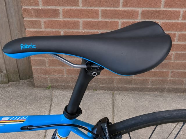 Fabric brand saddle on blue bike, black seatpost, non-drive side view against brick wall.