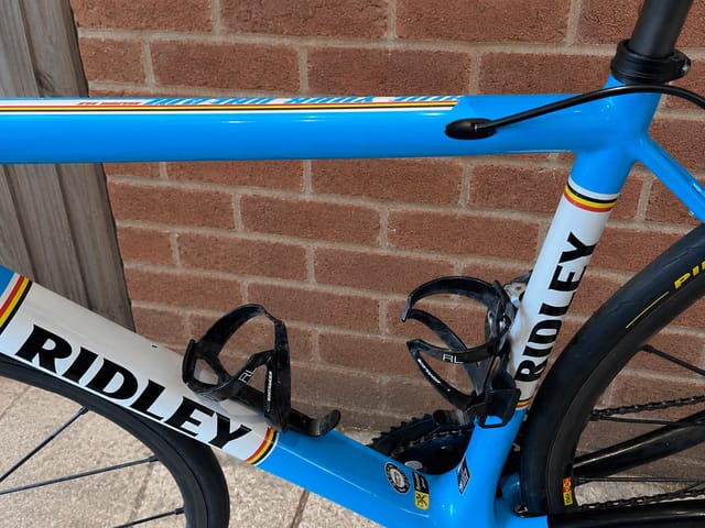 Blue and white Ridley road bike against a brick wall, drive side, showing frame detail with rainbow band and black drop bar shifters.